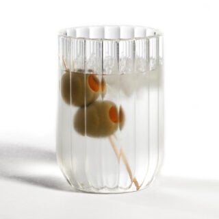 Minimalist white studio background with Fferrone Dearborn Water Glass in fluted borosilicate glass with a clear liquid and olive garnish | Kneen & Co