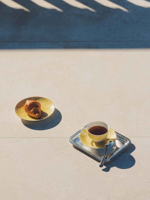 CHRISTOFLE-MALMAISON-RIVIERA-2026-RGB-HD-17-1200w-100KB Minimalist outdoor breakfast setting featuring the Christofle Malmaison Riviera Tea Cup and Bread Plate in yellow Limoges porcelain | Kneen & Co