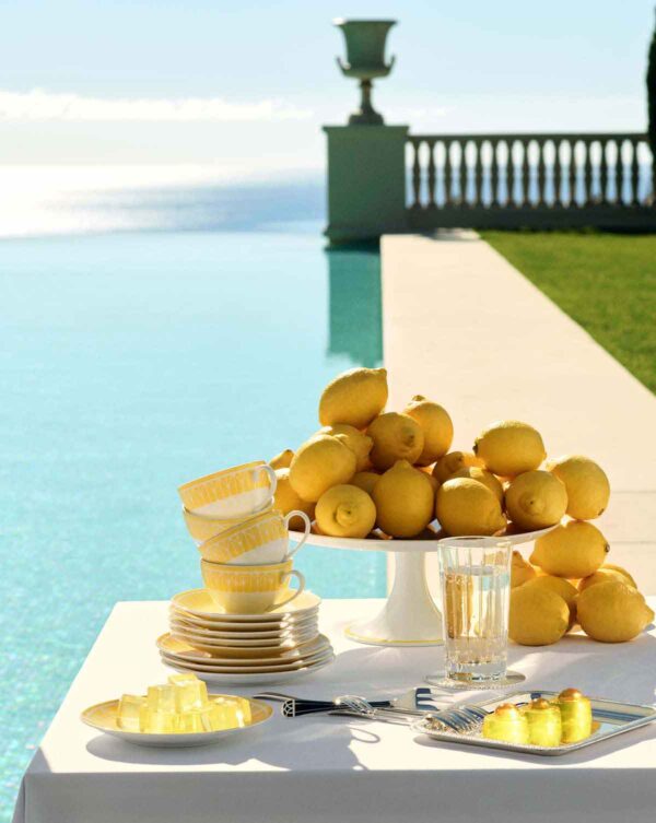 Scenic poolside table setting featuring Christofle Malmaison Riviera Tea Cups and Saucers in yellow Limoges porcelain alongside a porcelain cake stand | Kneen & Co