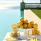 Scenic poolside table setting featuring Christofle Malmaison Riviera Tea Cups and Saucers in yellow Limoges porcelain alongside a porcelain cake stand | Kneen & Co