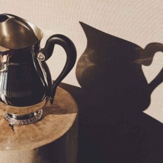 Artistic lifestyle setting featuring a Christofle Malmaison Silver-Plated Water Pitcher finished with a contrasting matte black ebony handle and intricate Empire-style leaf engravings on the base | Kneen & Co