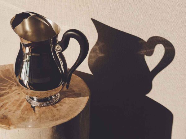 Artistic lifestyle setting featuring a Christofle Malmaison Silver-Plated Water Pitcher finished with a contrasting matte black ebony handle and intricate Empire-style leaf engravings on the base | Kneen & Co
