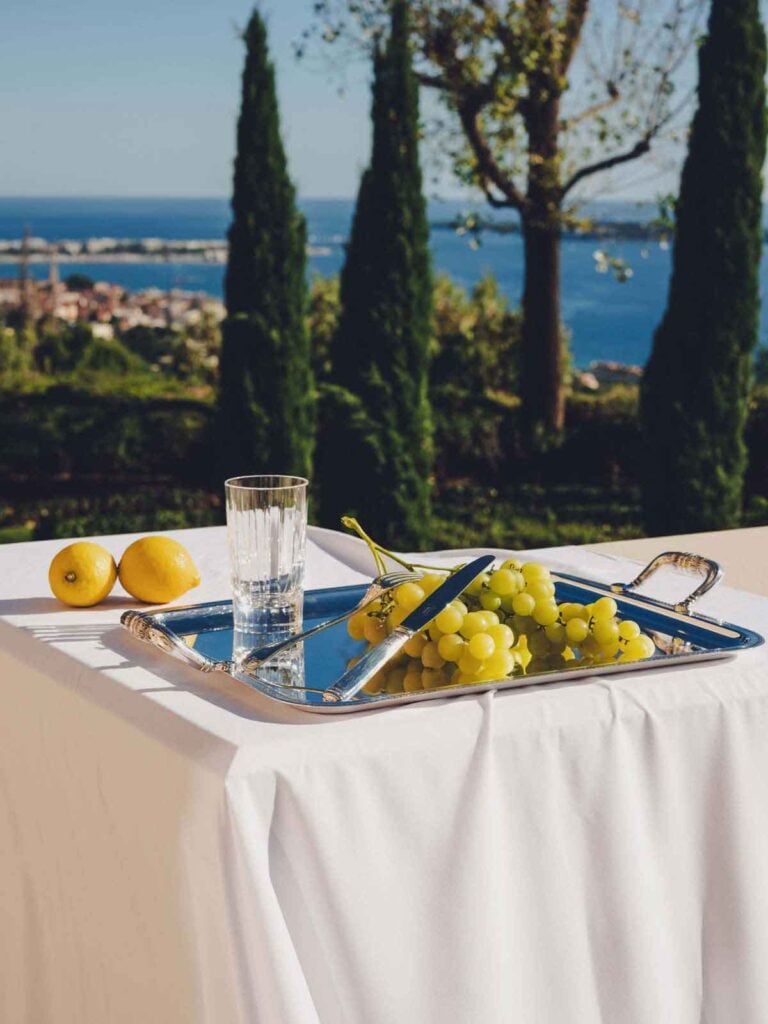 Sunlit outdoor Mediterranean setting featuring a handled silver-plated serving tray from the Christofle Malmaison collection and a crystal water tumbler | Kneen & Co