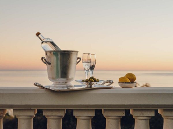 Serene coastal sunset setting featuring a Christofle Malmaison Champagne Bucket and handled tray in mirror-polished silver plate with signature Empire-style leaf borders and crystal flutes | Kneen & Co