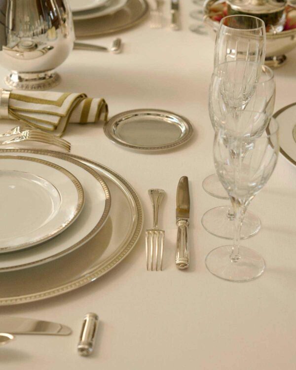 CHRISTOFLE-NewMalmaison-Mood-2026-RGB-HD-8-1200w-100KB Close-up of a formal place setting featuring Christofle Malmaison porcelain plates and silver-plated flatware | Kneen & Co