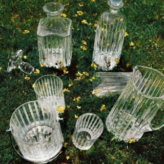 Baccarat Harmonie crystal barware, including a pitcher, decanters, tumblers, ice bucket, and cocktail shaker, displayed on grass among wildflowers, showcasing the collection's signature vertical cuts and contemporary design. | Kneen & Co.