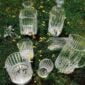 Baccarat Harmonie crystal barware, including a pitcher, decanters, tumblers, ice bucket, and cocktail shaker, displayed on grass among wildflowers, showcasing the collection's signature vertical cuts and contemporary design. | Kneen & Co.