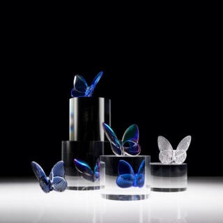 A collection of Baccarat Lucky Butterfly crystal sculptures in blue, iridescent, and clear crystal displayed on sculptural black and clear acrylic pedestals, highlighting Evelyne Julienne's graceful butterfly design and Baccarat's exceptional French crystal craftsmanship | Kneen & Co