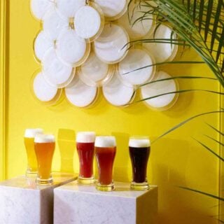 BACCARAT-2103547-Lifestyle-1200w-100KB Baccarat Oenologie Beer Highball glasses filled with a variety of craft beers, displayed on marble pedestals against a vibrant yellow backdrop, showcasing the collection's elegant curved silhouette and handcrafted French crystal design | Kneen & Co