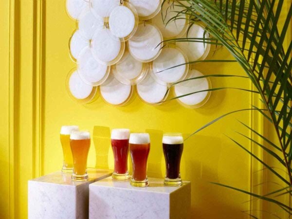 BACCARAT-2103547-Lifestyle-1200w-100KB Baccarat Oenologie Beer Highball glasses filled with a variety of craft beers, displayed on marble pedestals against a vibrant yellow backdrop, showcasing the collection's elegant curved silhouette and handcrafted French crystal design | Kneen & Co