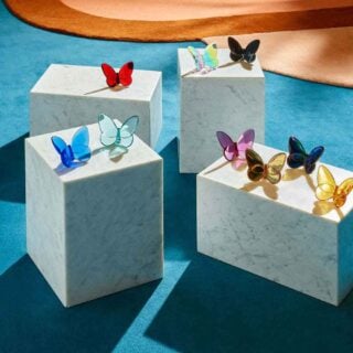 Baccarat Lucky Butterflies crystal collection displayed in vibrant colors atop white marble pedestals, showcasing decorative butterfly sculptures in clear, blue, red, pink, amber, iridescent, and black crystal | Kneen & Co