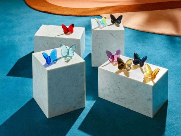 Baccarat Lucky Butterflies crystal collection displayed in vibrant colors atop white marble pedestals, showcasing decorative butterfly sculptures in clear, blue, red, pink, amber, iridescent, and black crystal | Kneen & Co