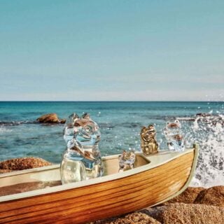 Baccarat Maneki Neko crystal lucky cat figurines displayed in a wooden boat on a rocky shoreline, featuring clear and gold crystal designs against a seaside backdrop with ocean waves | Kneen & Co