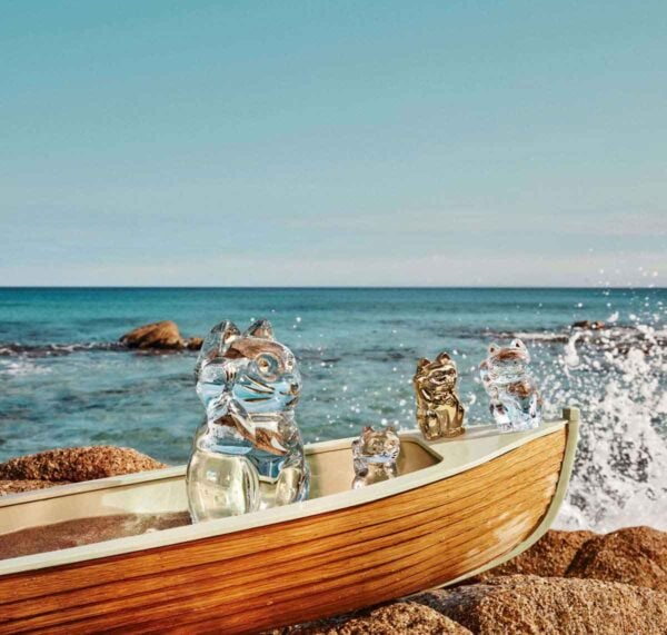 BACCARAT-2803413-1200w-100KB Baccarat Maneki Neko crystal lucky cat figurines displayed in a wooden boat on a rocky shoreline, featuring clear and gold crystal designs against a seaside backdrop with ocean waves | Kneen & Co