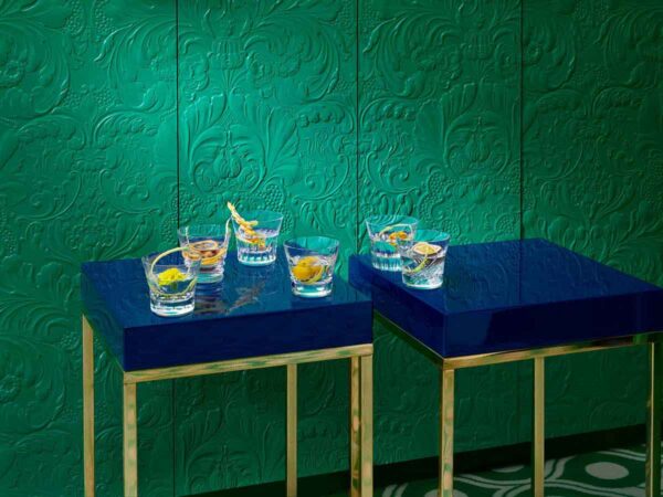 Baccarat Everyday Classic crystal tumblers displayed on deep blue lacquer tables against an emerald green textured wall, each glass featuring a unique cut pattern and garnished cocktails that showcase the collection's versatile design | Kneen & Co