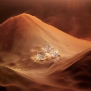 BACCARAT-2811294-Lifestyle-1200w-100KB Baccarat Masséna crystal highball glasses and tumbler displayed on sculpted sand dunes, highlighting the collection's signature deep bevel cuts and brilliant light-reflecting crystal | Kneen & Co