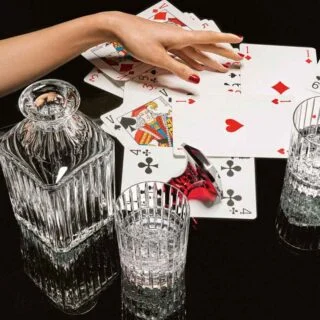 BACCARAT-2811298-1845261-2811293-2811299-2813210-1704365-Lifestyle-1200w-100KB Baccarat Harmonie crystal tumblers and matching whiskey decanter displayed with playing cards, showcasing the collection's signature vertical fluting for refined whisky service and cocktails | Kneen & Co