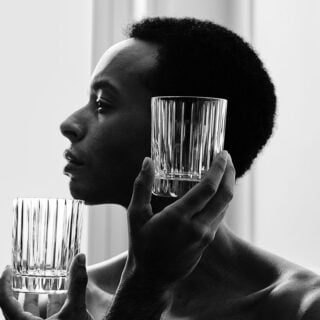 BACCARAT-2811298-1845261-2811293-2811299-2813210-Lifestyle-1-1200w-100KB Two Baccarat Harmonie crystal tumblers featuring the collection's signature vertical fluting held against a portrait backdrop, highlighting the clean architectural lines and timeless design | Kneen & Co