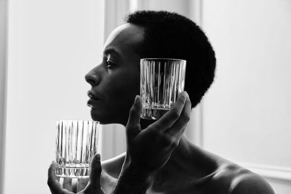 BACCARAT-2811298-1845261-2811293-2811299-2813210-Lifestyle-1-1200w-100KB Two Baccarat Harmonie crystal tumblers featuring the collection's signature vertical fluting held against a portrait backdrop, highlighting the clean architectural lines and timeless design | Kneen & Co