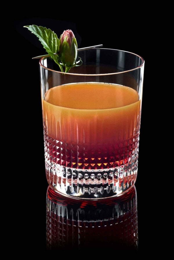 BACCARAT-2811580-Lifestlye-1200w-100KB Baccarat Nancy crystal tumbler filled with a layered cocktail and garnished with a fresh hibiscus bud, showcasing the collection's signature diamond-cut crystal pattern | Kneen & Co