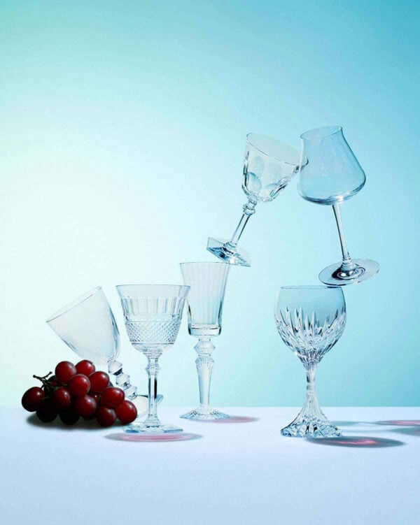 BACCARAT-2812727-Lifestyle-1-1200w-100KB Baccarat crystal stemware and barware arranged in a contemporary still life with red grapes against a soft blue background, showcasing the Maison's iconic French crystal craftsmanship | Kneen & Co