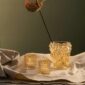 Earthy studio setting featuring the Lalique Bacchantes Small Vase in gold-lustre crystal, next to two small glowing votives | Kneen & Co